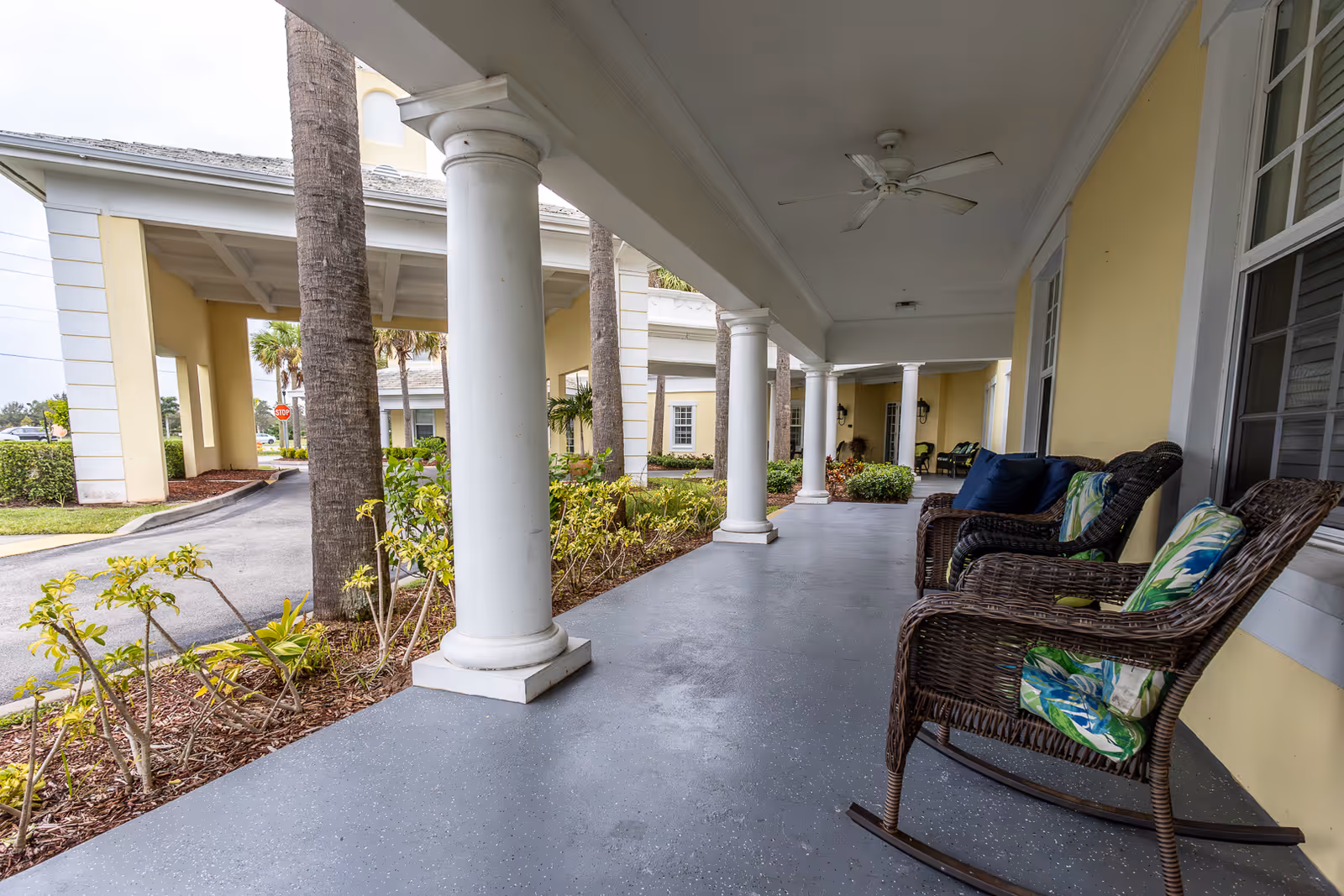 Covered front porch of a yellow building with white columns, ceiling fans, and wicker rocking chairs with cushions.