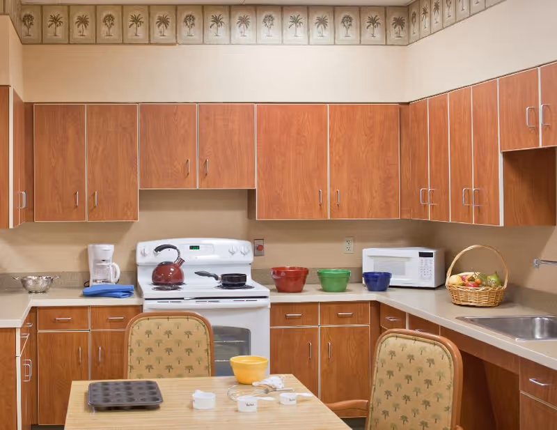 A kitchen area with wooden cabinets, a white stove with a red kettle and a pan on it, a white microwave, a coffee maker, a basket of fruit, and a table with baking items including a muffin tray, bowls, and measuring cups. Two chairs with palm tree patterned upholstery are positioned at the table.