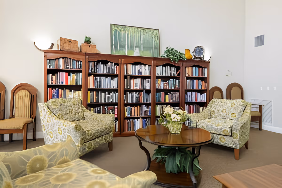 Cozy sitting area with patterned armchairs around a coffee table in front of a large wooden bookshelf filled with books.