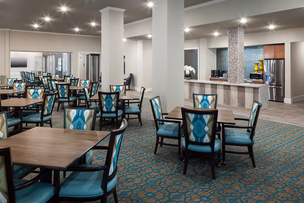 A spacious dining room with multiple wooden tables and chairs featuring blue and patterned upholstery. The room has a patterned carpet in shades of blue and orange, recessed ceiling lights, and a kitchen area with a refrigerator, microwave, and decorative vases in the background.