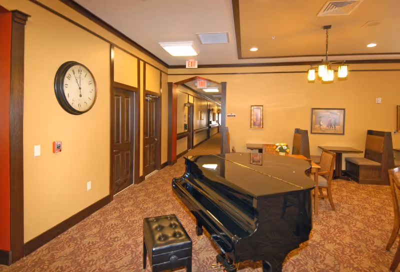 Interior view of a common area in a retirement center featuring a black grand piano with a matching bench in the foreground. The room has warm beige walls with dark wood trim, a patterned carpet, and several wooden booths and tables with chairs. There are framed pictures on the walls and ceiling lights providing illumination. A hallway with exit signs is visible in the background.