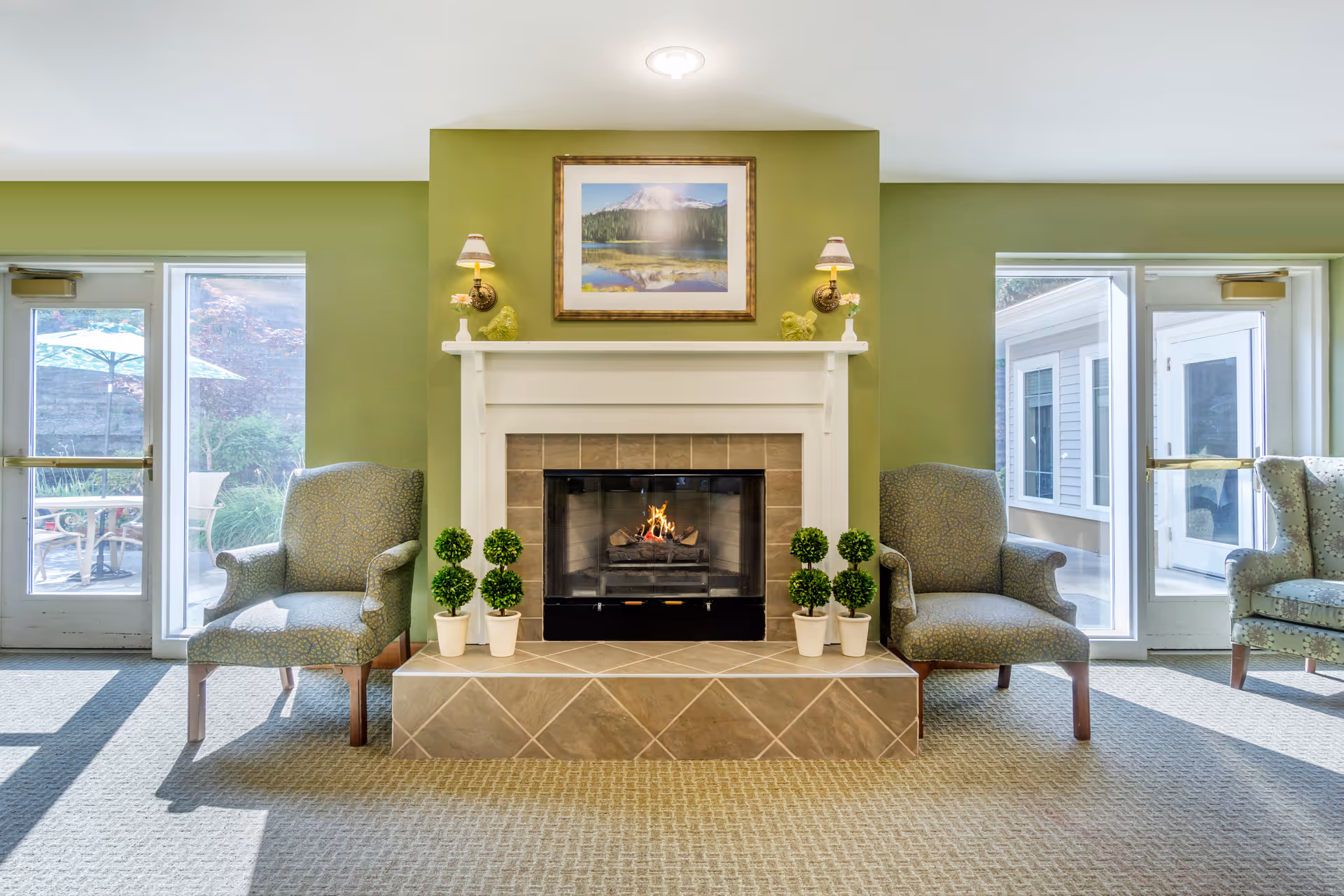 A cozy living room area with a lit fireplace centered on a green wall. Above the fireplace is a framed picture of a mountain and lake. On either side of the fireplace are two matching armchairs with patterned upholstery. Small potted topiary plants are placed symmetrically on the tiled hearth. There are glass doors on both sides of the fireplace leading to an outdoor patio area with umbrellas and chairs visible outside.