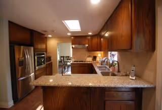 Interior view of a kitchen with wooden cabinets, granite countertops, a stainless steel refrigerator, built-in oven, and a sink. The kitchen opens into a dining area with a table and chairs visible in the background.
