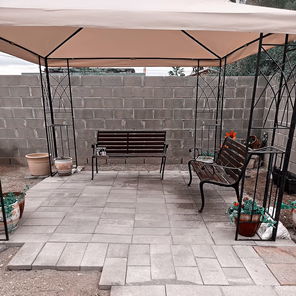 Outdoor seating area with two metal and wood benches under a beige canopy. The area is paved with stone tiles and surrounded by a concrete block wall. Several potted plants are placed around the seating area.