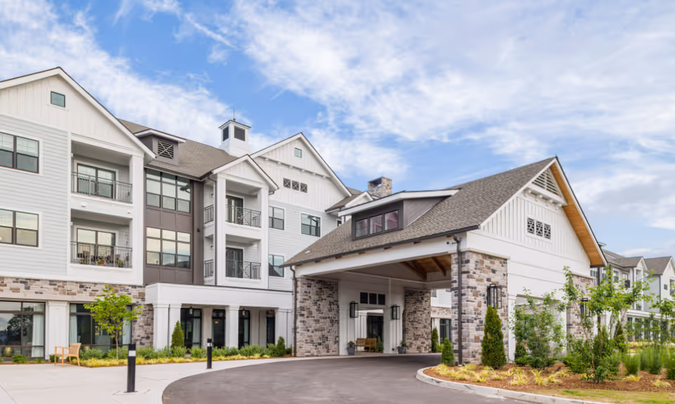 Exterior view of a multi-story senior living facility with a covered entrance, stone and white siding facade, landscaped greenery, and a partly cloudy sky.