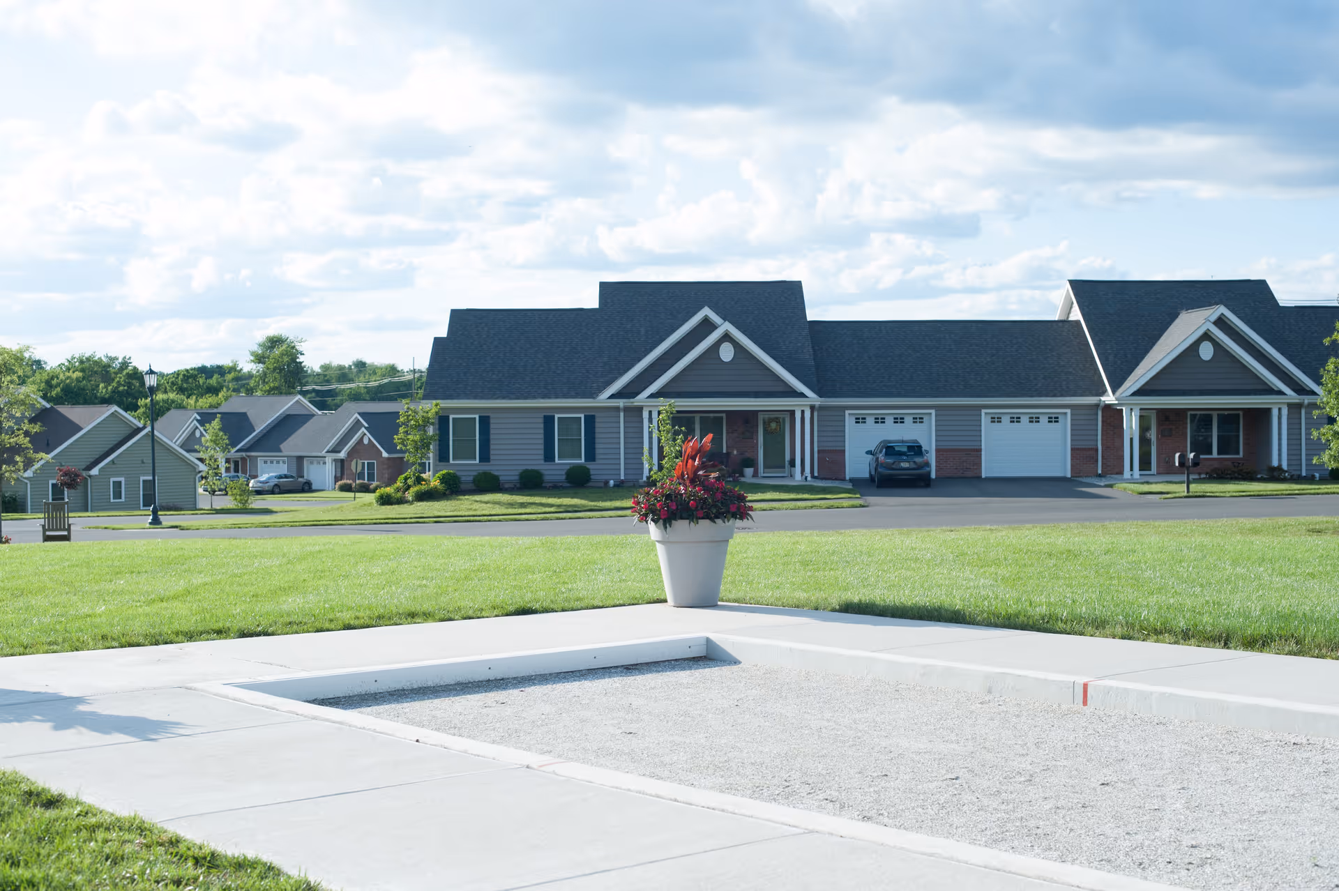 Row of single-story residential units with garages set behind a grassy lawn and a planter in the foreground.