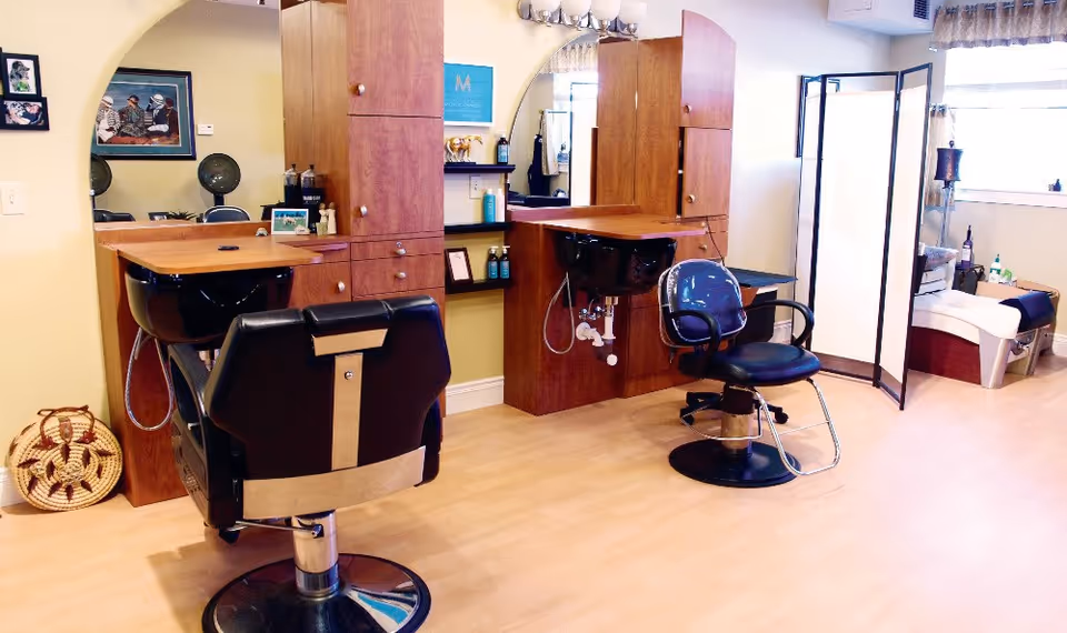 Interior of a senior living facility's hair salon area with two black salon chairs facing wooden cabinets with sinks for hair washing. The room has light-colored wooden flooring, a folding privacy screen, and a window with curtains letting in natural light.