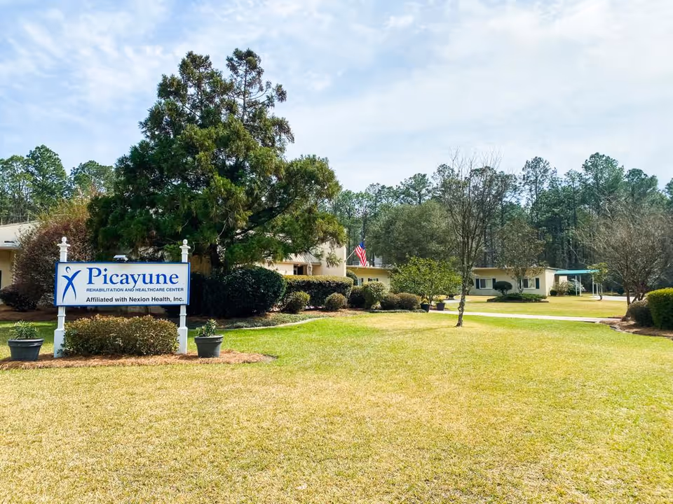 Exterior view of Picayune Rehabilitation and Healthcare Center showing a large grassy lawn with trees and bushes. A sign with the facility name is prominently displayed in the foreground. The building is visible in the background under a partly cloudy sky.