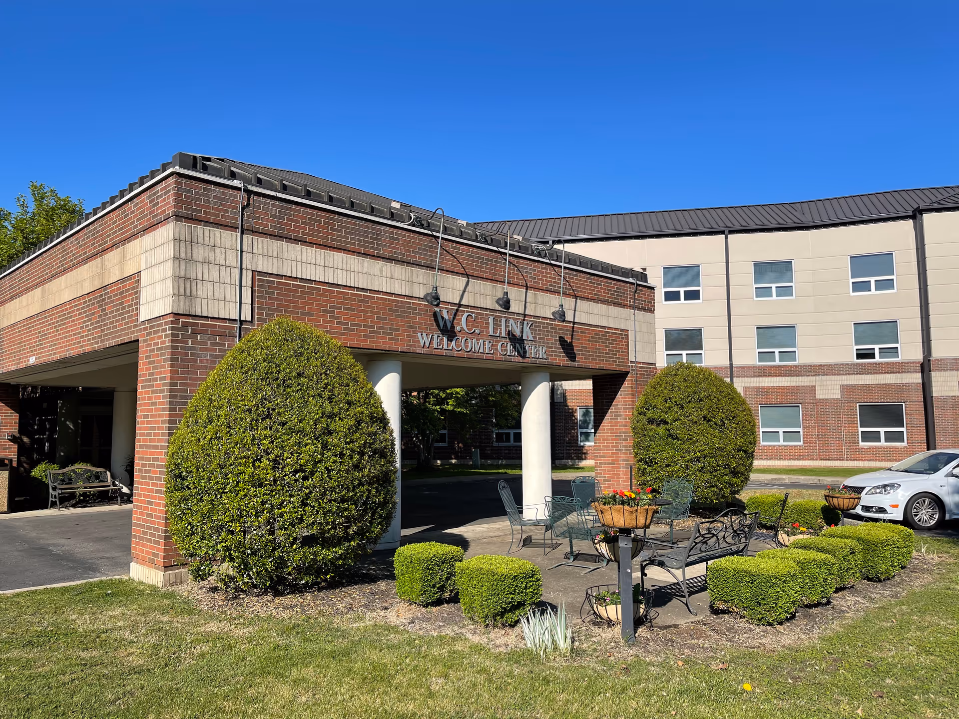 Brick building front with a covered portico labeled "Welcome Center", trimmed shrubs, outdoor seating and a parked car under a clear blue sky.