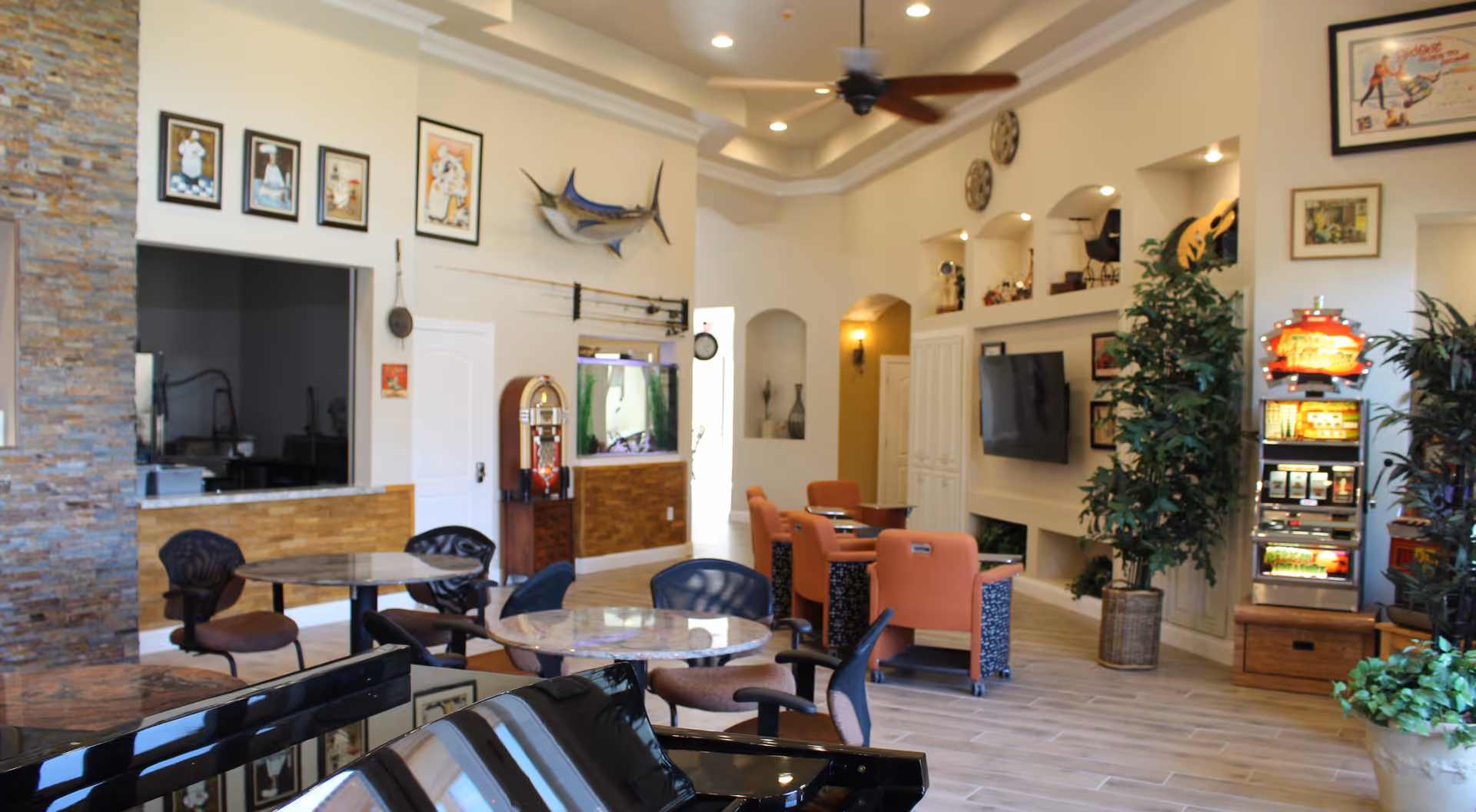 Spacious assisted living common area with round tables and chairs, a piano in the foreground, wall-mounted TV, plants and decorative wall niches.