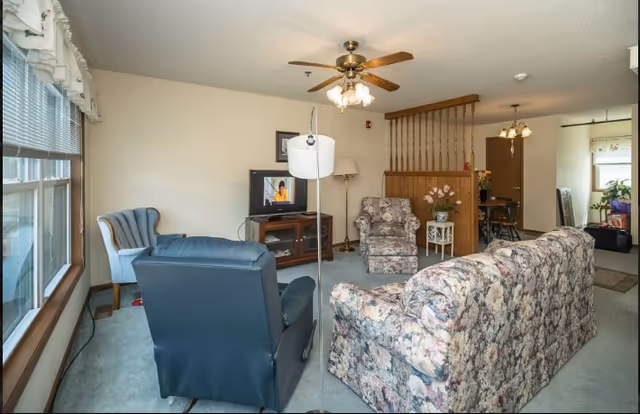 A cozy living room with floral patterned sofa and armchair, a blue recliner, and a light blue wingback chair near a large window with blinds. A wooden TV stand holds a television and a lamp, and a ceiling fan with lights hangs above. The room has light-colored walls and carpeted floor, with a small table holding a vase of flowers near a wooden partition separating the living area from a dining space in the background.
