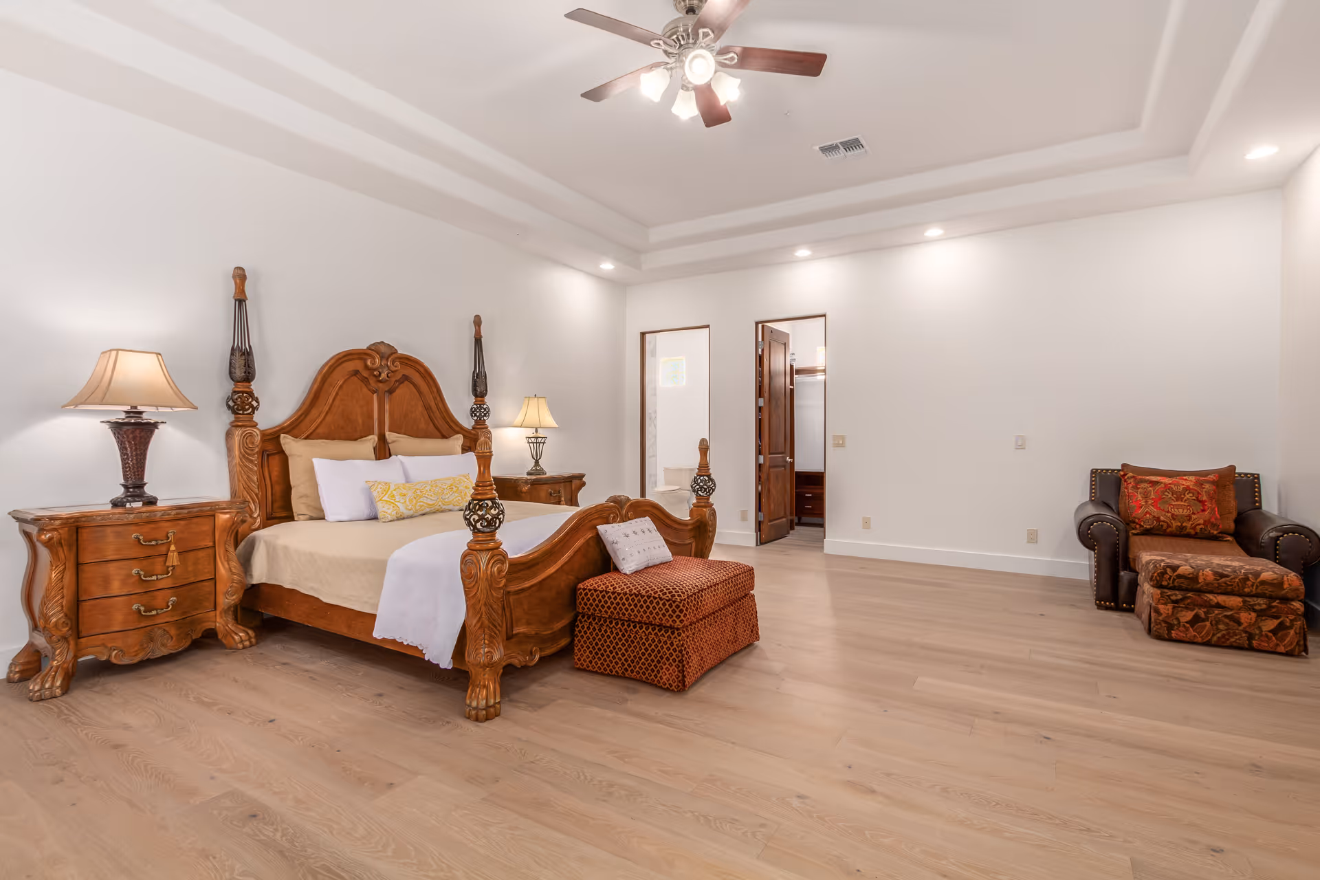 Spacious bedroom with an ornate wooden bed, matching nightstands and lamps, an ottoman, and an upholstered armchair on light wood floors.
