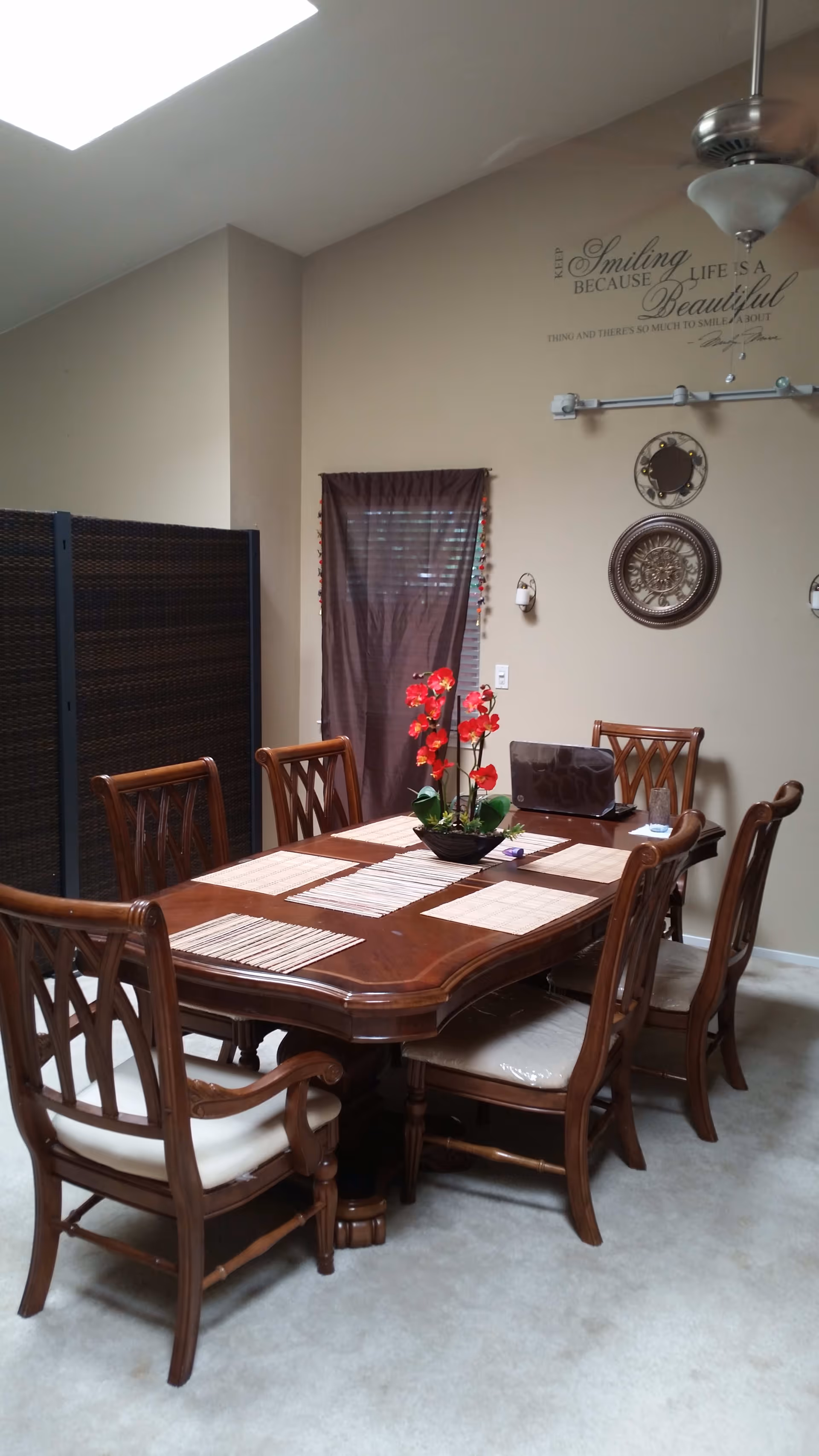 A dining room with a wooden dining table and six matching chairs with cushioned seats. The table has placemats and a centerpiece with red flowers. There is a brown curtain covering a window, decorative wall hangings including a round mirror and a sunburst design, and a ceiling fan with a light fixture above the table.