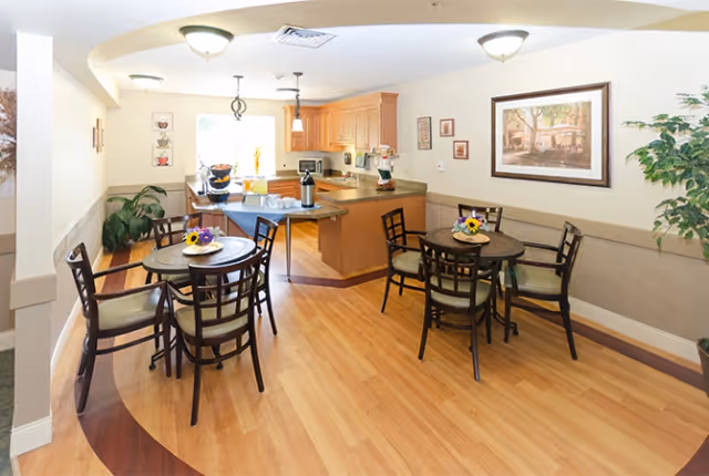 Bright communal dining area with round tables and chairs opening to a kitchen with wood cabinets and a serving counter.
