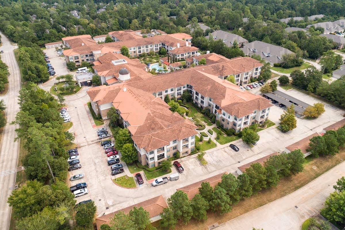 Aerial view of Conservatory At Alden Bridge, a large senior living facility with multiple connected buildings featuring red-tiled roofs, surrounded by trees and parking lots with cars. The facility is located in a green, suburban area with nearby residential houses.