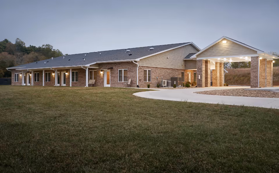 Single-story brick retirement community building with a covered porte-cochère and a large grassy front lawn at dusk.
