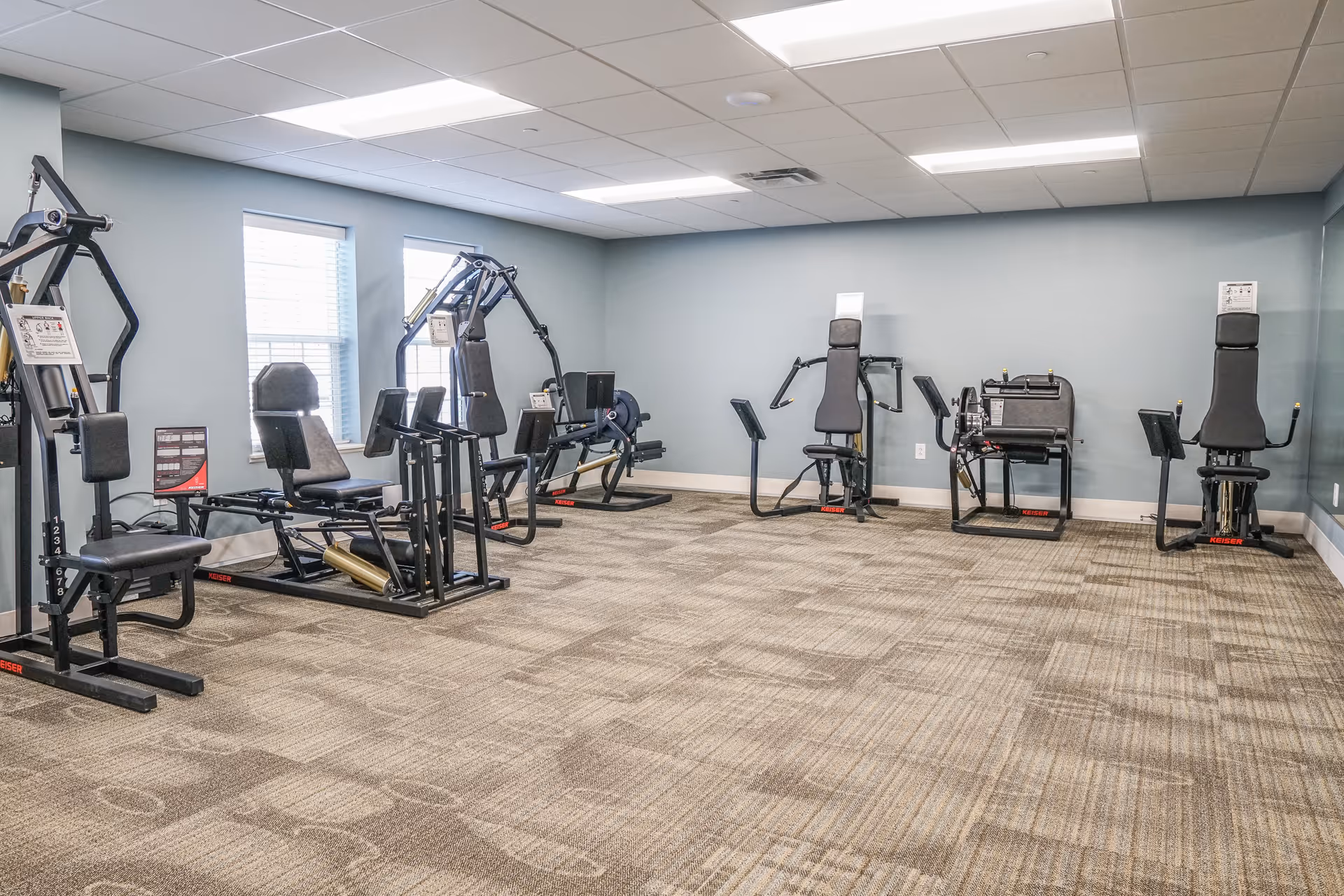A fitness room with multiple exercise machines arranged along the walls. The room has light blue walls, two windows with blinds, and a patterned carpet floor. The ceiling has recessed lighting panels.