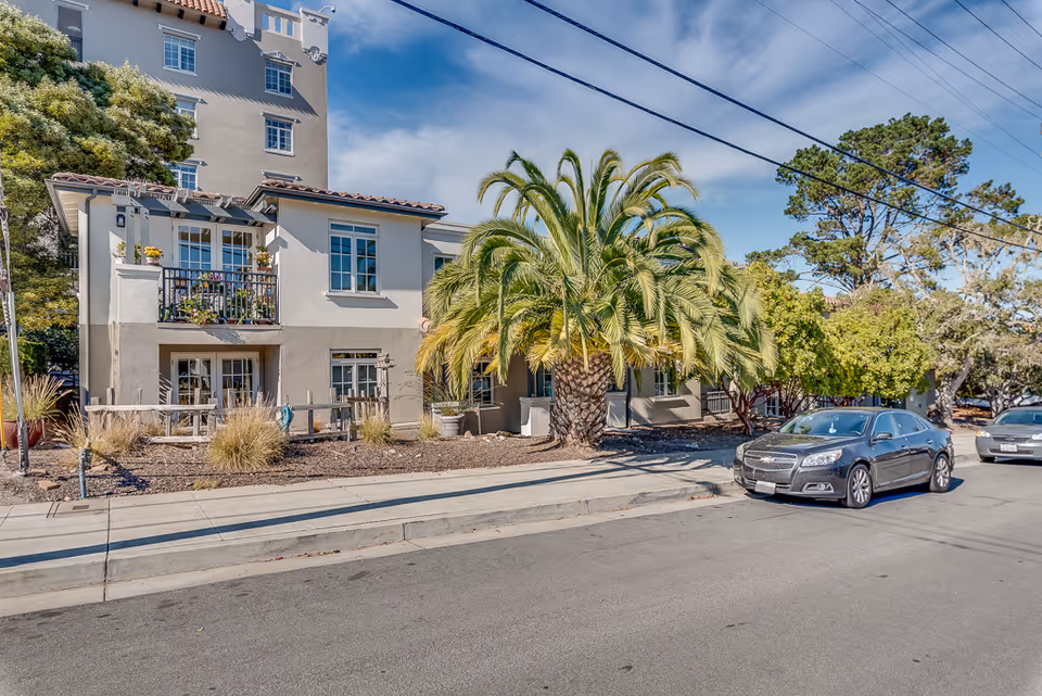 Exterior view of a senior living facility building with beige walls and multiple windows. There is a large palm tree and other greenery in front of the building, along with a sidewalk and parked cars on the street. The sky is clear with some clouds.