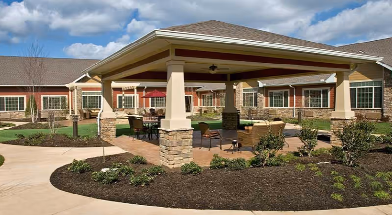 Outdoor covered seating area with stone pillars and a roof, surrounded by a garden with shrubs and a paved walkway. The background shows a single-story building with multiple windows under a partly cloudy sky.