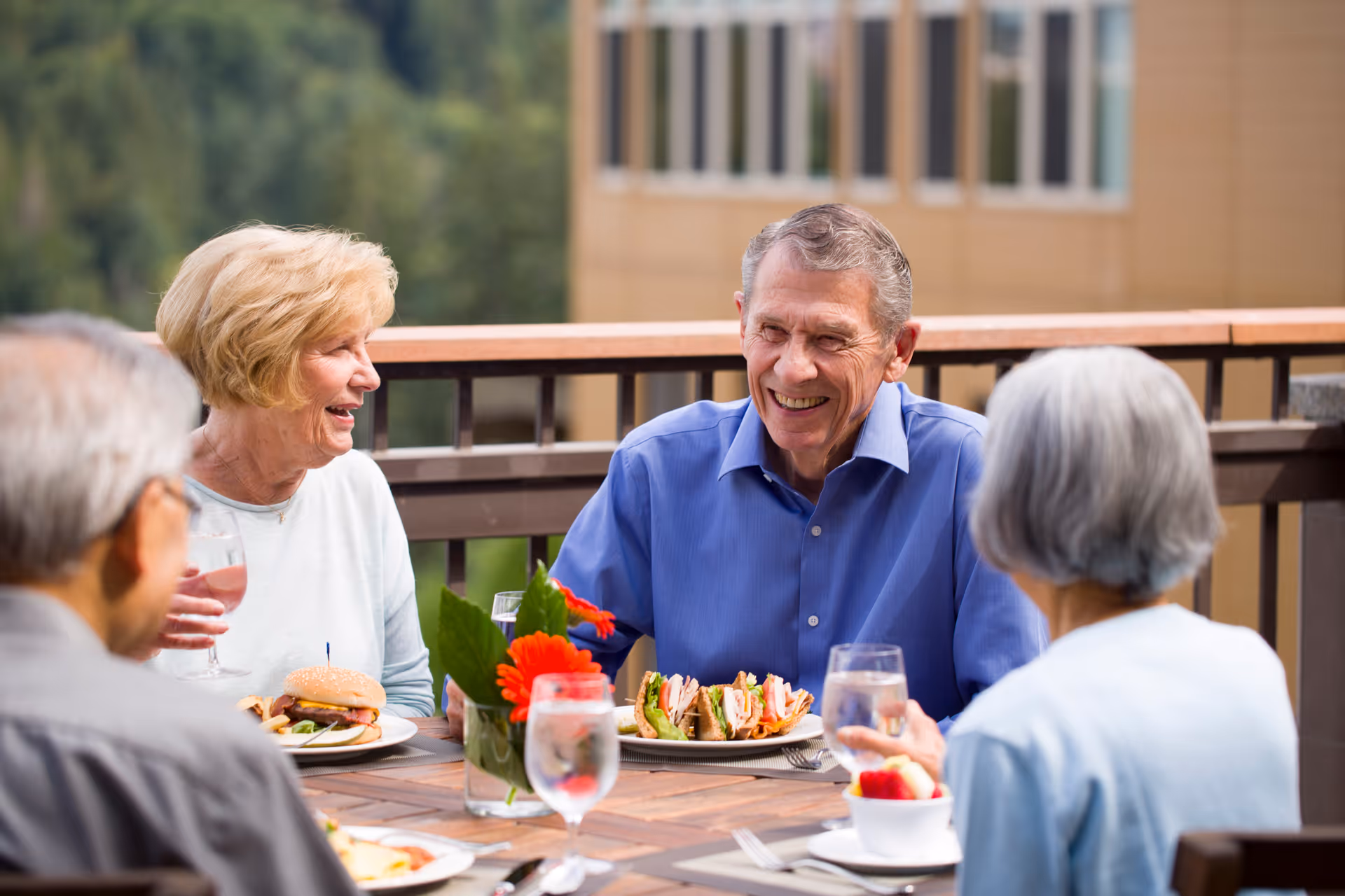Four elderly people sitting around a wooden table outdoors, enjoying a meal together. The table has plates with sandwiches and glasses of water, and there is a small vase with red flowers in the center. They appear to be engaged in a pleasant conversation on a balcony or patio with a building and greenery in the background.