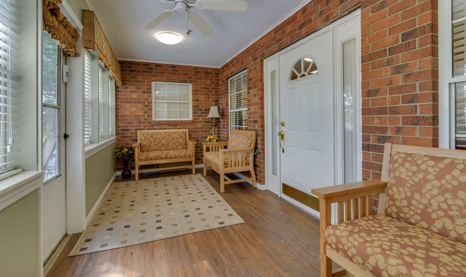 A cozy enclosed porch area with brick walls, a white door with glass panels, and several wooden chairs with patterned cushions. The floor is wood with a beige area rug, and there are windows with blinds and valances letting in natural light. A ceiling fan and a lamp on a small table add to the comfortable atmosphere.