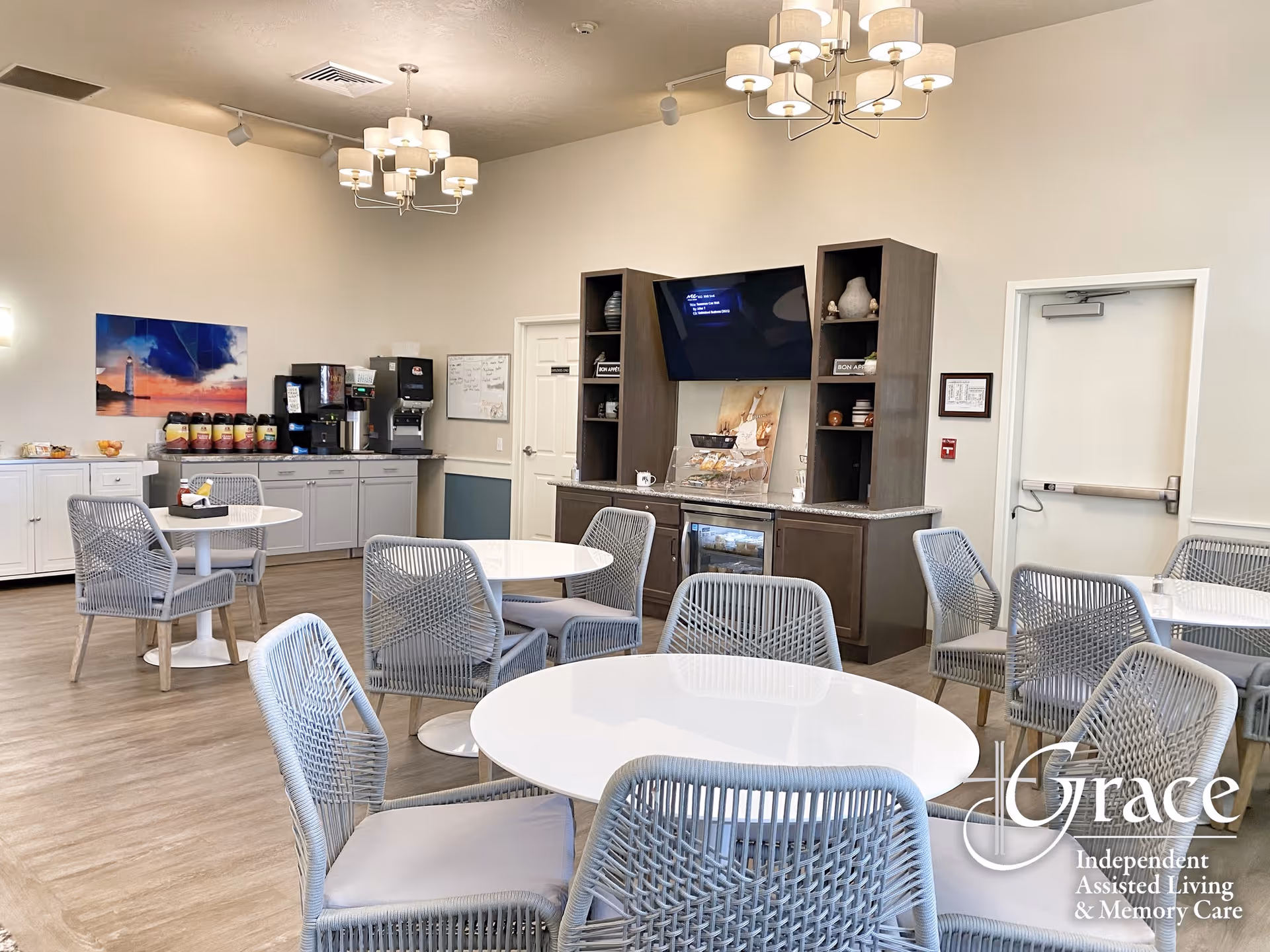 Bright communal dining area with round white tables, woven chairs, a coffee station and a TV on the wall.