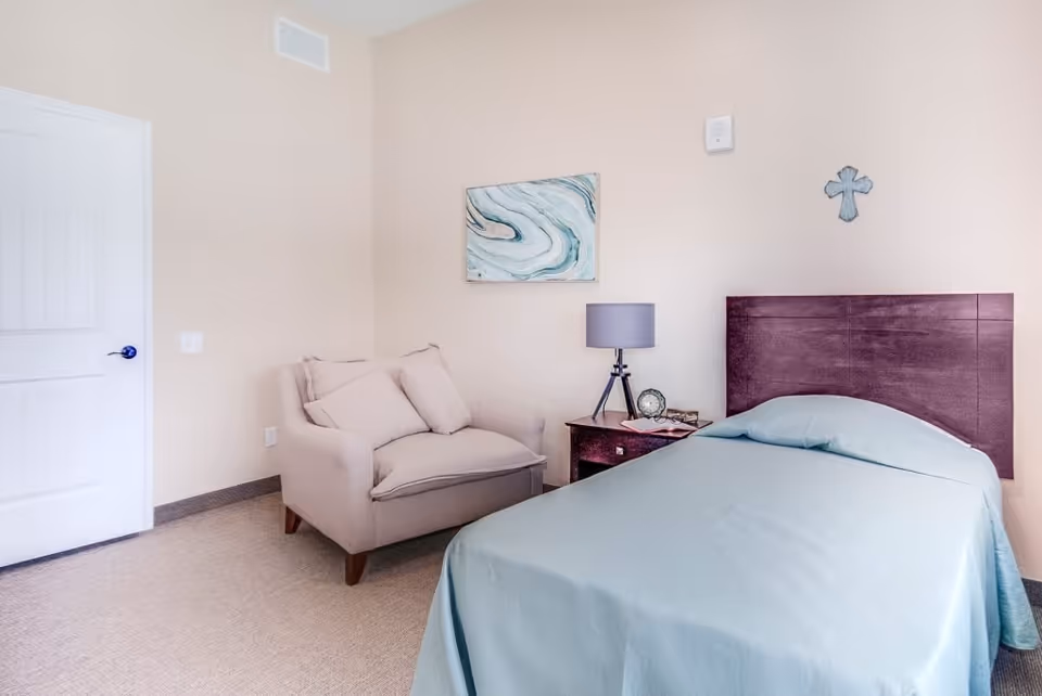 A simple bedroom with a single bed covered in a light blue bedspread, a wooden headboard, a small wooden nightstand with a lamp, clock, and glasses, a beige armchair with two cushions, a door, and a wall decorated with a blue and white abstract painting and a small cross.