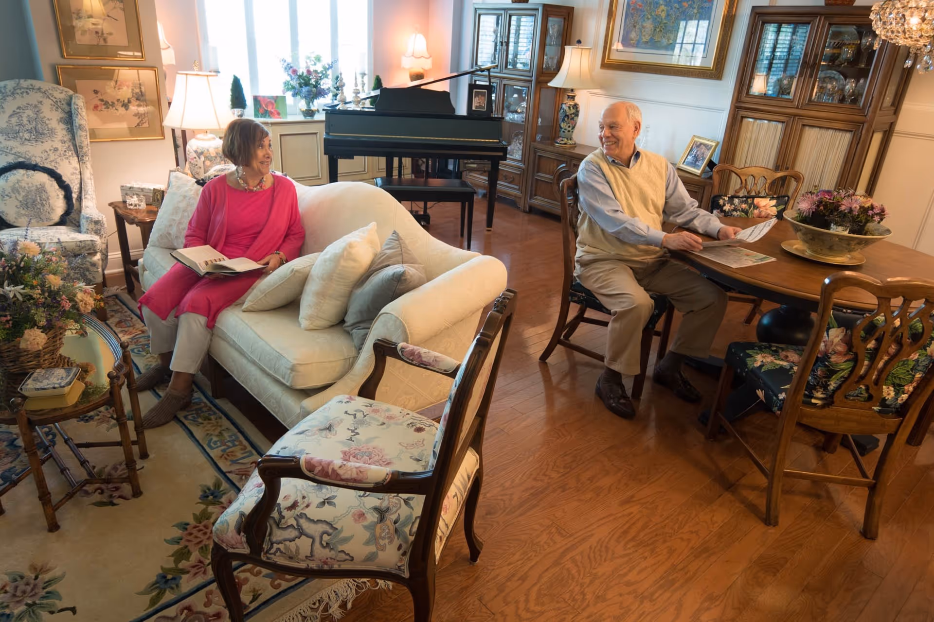 An elderly woman in a pink outfit sits on a cream-colored sofa holding a book, smiling at an elderly man who is seated at a wooden dining table reading a newspaper. The room is warmly decorated with floral-patterned chairs, a large area rug, a grand piano near the window, and wooden cabinets displaying decorative items.