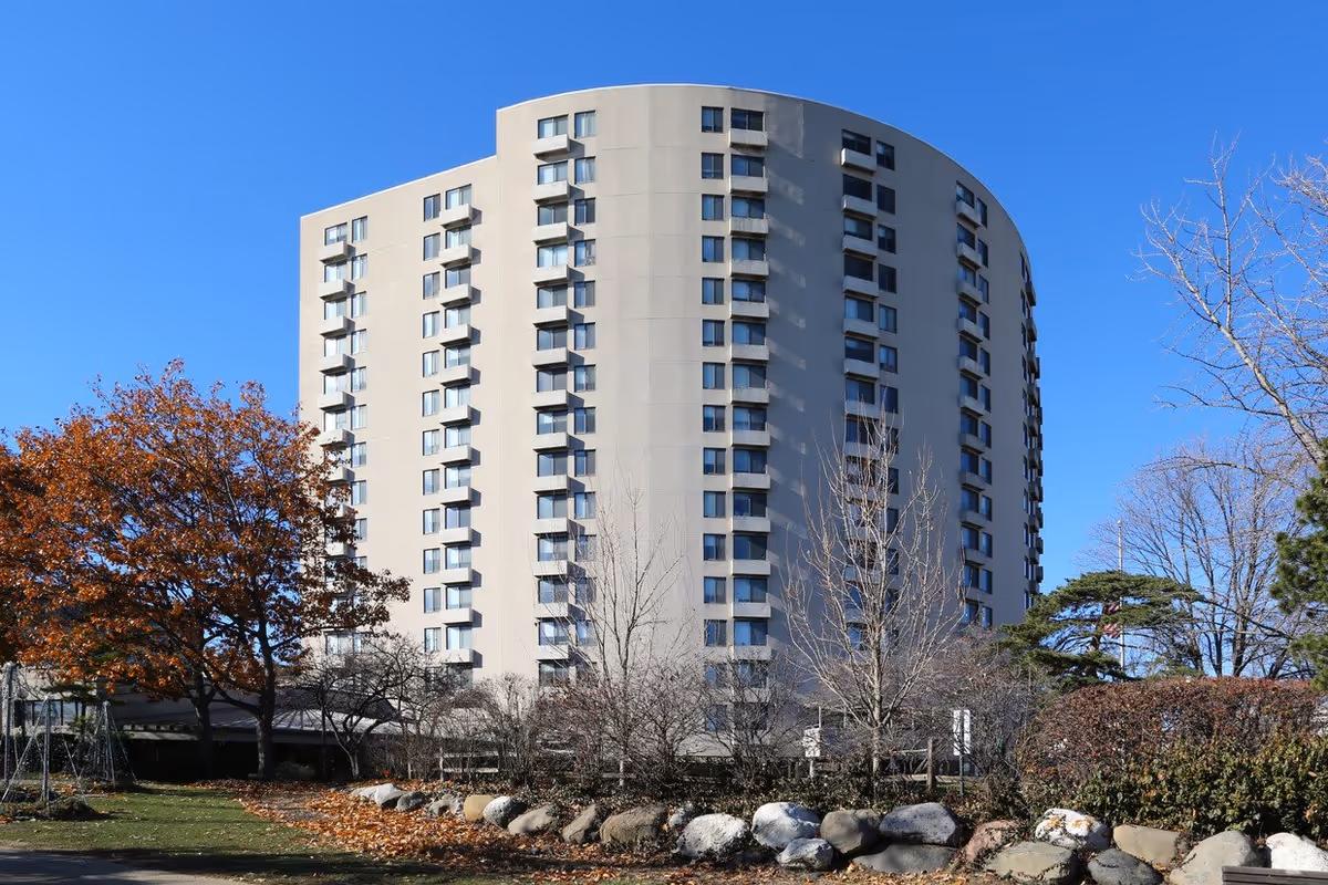 Tall, curved multi-story residential building with many windows, surrounded by trees with autumn foliage and a clear blue sky.