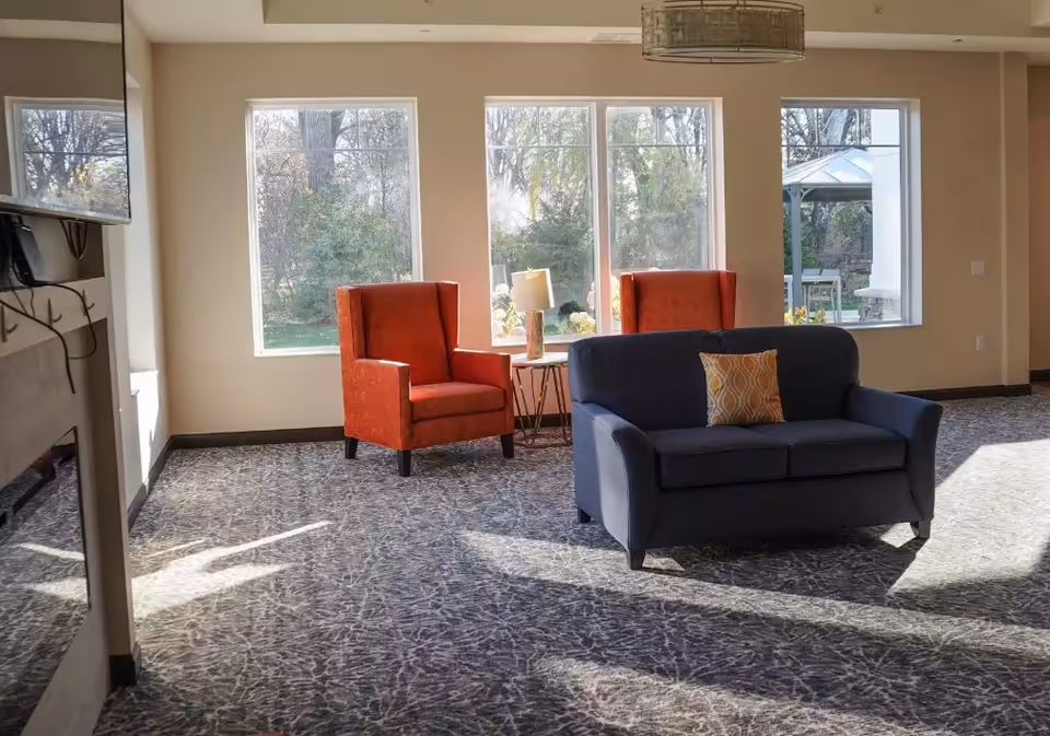 A bright living room area with three large windows letting in natural light. The room features a dark blue loveseat with a patterned throw pillow, two orange armchairs, and a small round table with a lamp between the armchairs. Outside the windows, trees and a gazebo are visible.