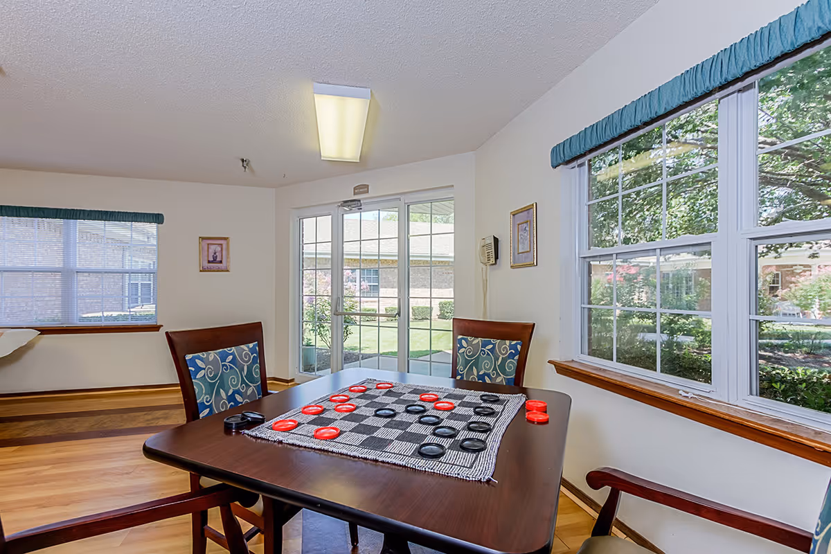 A bright room with large windows showing a garden outside. Inside, there is a wooden table with a checkers game set up, surrounded by chairs with patterned cushions. The room has light-colored walls, wooden flooring, and a glass door leading outside.