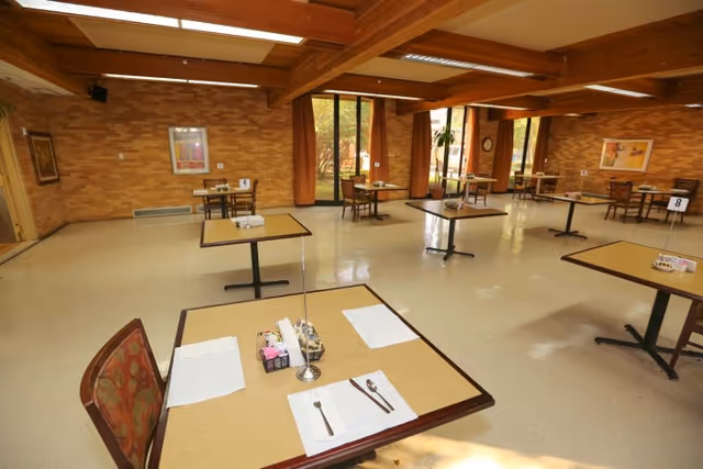 A spacious dining room with several square tables set with placemats, utensils, and condiment holders. The room has large windows with orange curtains, brick walls, and wooden ceiling beams. Each table has a number stand, and the floor is a light-colored linoleum.
