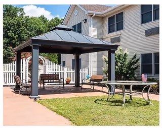 Outdoor patio area with a metal gazebo covering benches, a glass-top table with chairs on a grassy lawn, and a multi-story beige building in the background under a partly cloudy sky.