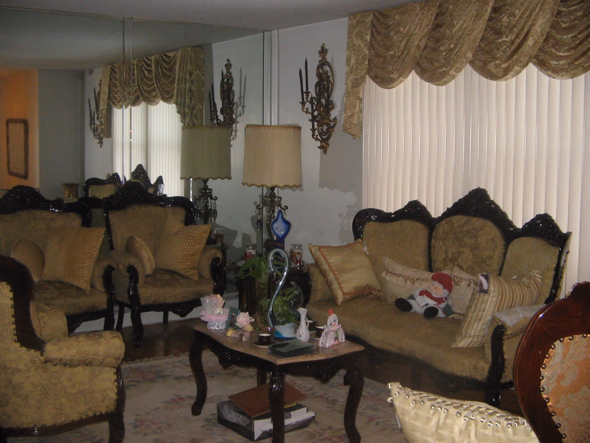 A traditional living room with ornate dark wood-framed upholstered sofas and armchairs in a beige patterned fabric. The room features a glass-topped coffee table with decorative items, a large mirror on one wall, vertical blinds covering the window, and a pair of vintage-style wall sconces and a table lamp with a pleated shade. There are several throw pillows and a small stuffed Santa Claus on one of the sofas.