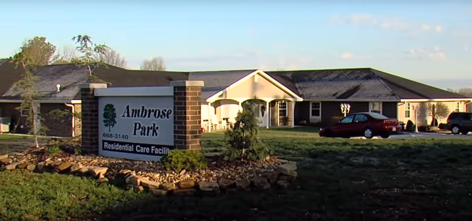 Exterior view of Ambrose Park Residential Care Facility showing a single-story brick building with a pitched roof, a lawn with some small trees and shrubs, a parking area with cars, and a large sign in front displaying the facility's name and phone number.