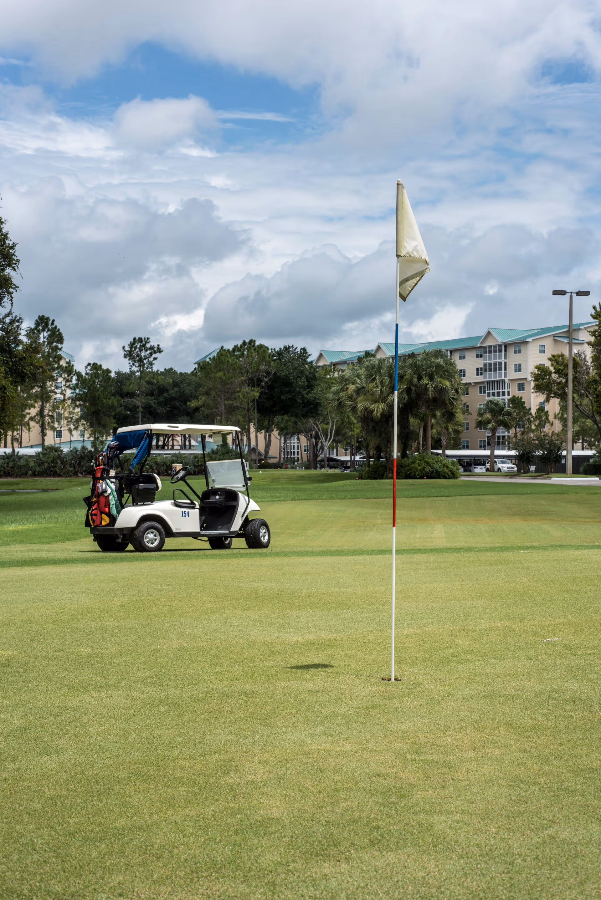 A golf course putting green with a flagstick in the hole. A white golf cart with golf bags is parked on the grass nearby. In the background, there are trees and a multi-story building with a teal roof under a partly cloudy sky.