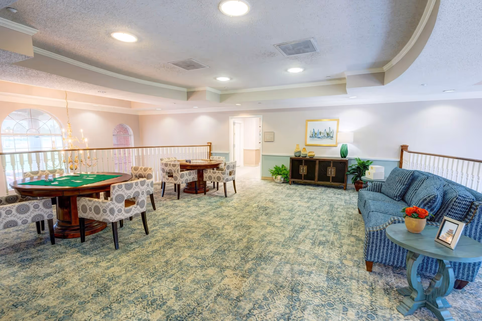 A spacious, well-lit common area with patterned carpet flooring and light-colored walls. The room features two round wooden tables with patterned upholstered chairs around them, suitable for games or socializing. A blue upholstered sofa is positioned near a small round side table with a potted plant and a framed photo. There is a wooden cabinet against the far wall with decorative items and a framed picture above it. Large windows with arched tops allow natural light to enter the space.