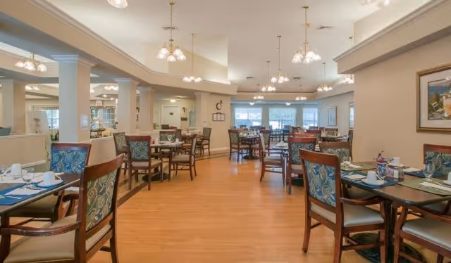 A spacious dining room with multiple wooden tables and chairs arranged neatly. Each table is set with placemats, cups, and utensils. The room has warm lighting from ceiling fixtures and large windows letting in natural light. The walls are decorated with framed artwork and the floor is wooden.