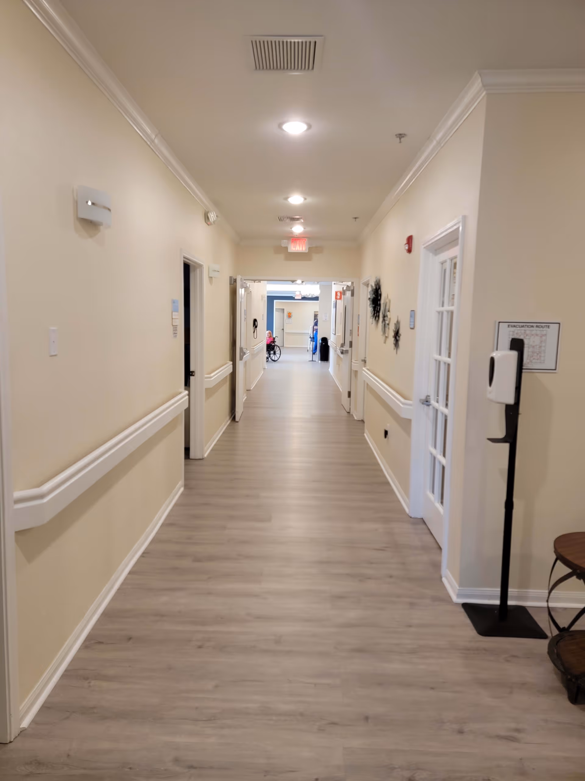 A long, well-lit hallway in a senior living facility with light-colored walls and wood flooring. There are handrails on both sides of the hallway, several doors leading to rooms, and a hand sanitizer dispenser on a stand near the right wall. At the far end, a person in a wheelchair and another person are visible.