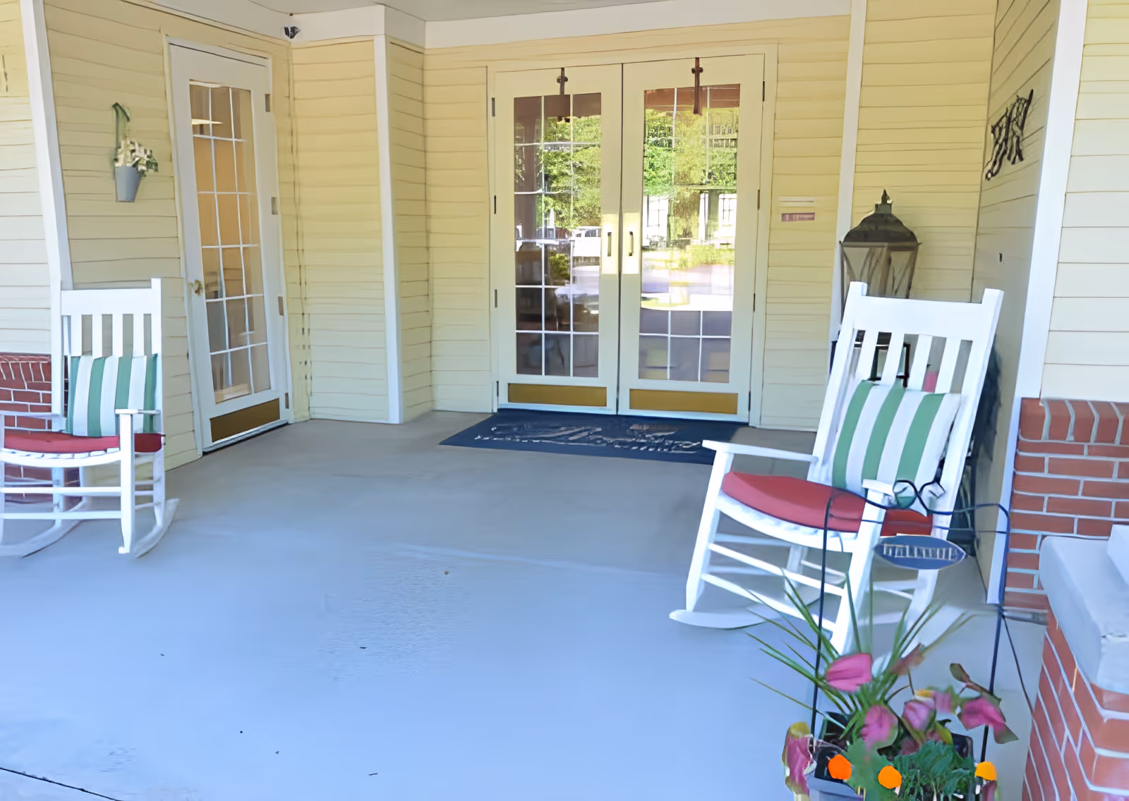 Covered porch area with two white rocking chairs featuring red cushions and green striped pillows, a potted plant with pink and orange flowers, and double glass doors leading inside a building with yellow siding and brick accents.