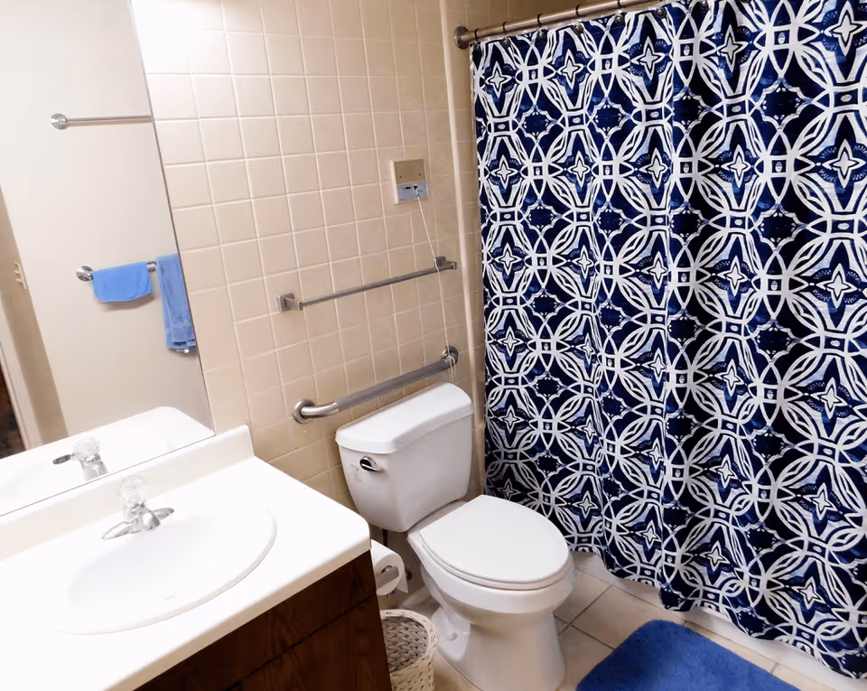 Bathroom with a white sink and countertop, a toilet, and a shower with a blue and white patterned curtain. There are beige tiles on the wall behind the toilet, a grab bar next to the toilet, and a blue bath mat on the floor. A mirror and towel rack with a blue towel are visible on the left side.