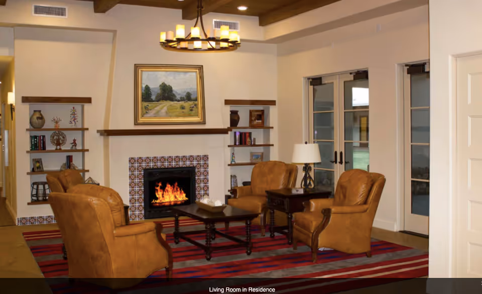A cozy living room with four brown leather armchairs arranged around a dark wooden coffee table on a red and gray striped rug. There is a fireplace with a decorative tile surround and a landscape painting above it. Built-in shelves with books and decorative items flank the fireplace. A table lamp sits on a side table next to one of the armchairs. Double glass doors lead to an outdoor area.
