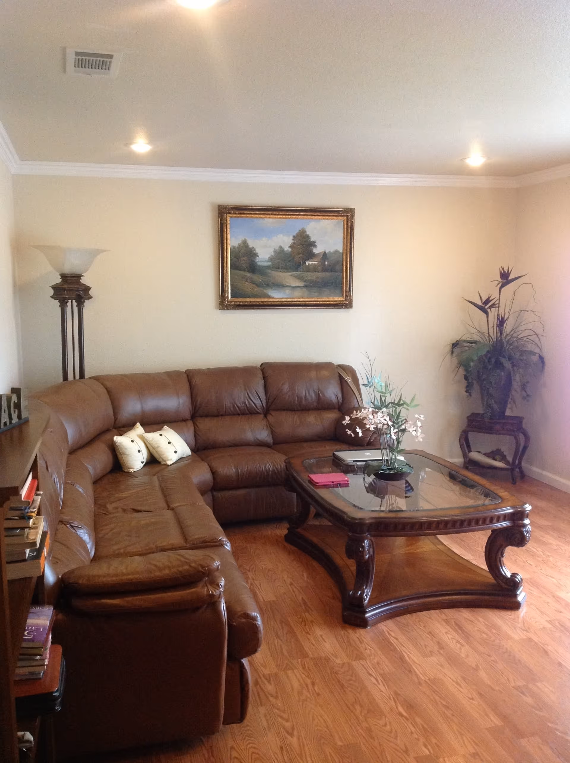 A cozy living room with a large brown leather sectional sofa, a wooden coffee table with a glass top, and a decorative plant on the right side. There is a floor lamp in the corner and a framed landscape painting on the beige wall above the sofa. The floor is wooden, and there are a few books on a shelf to the left.