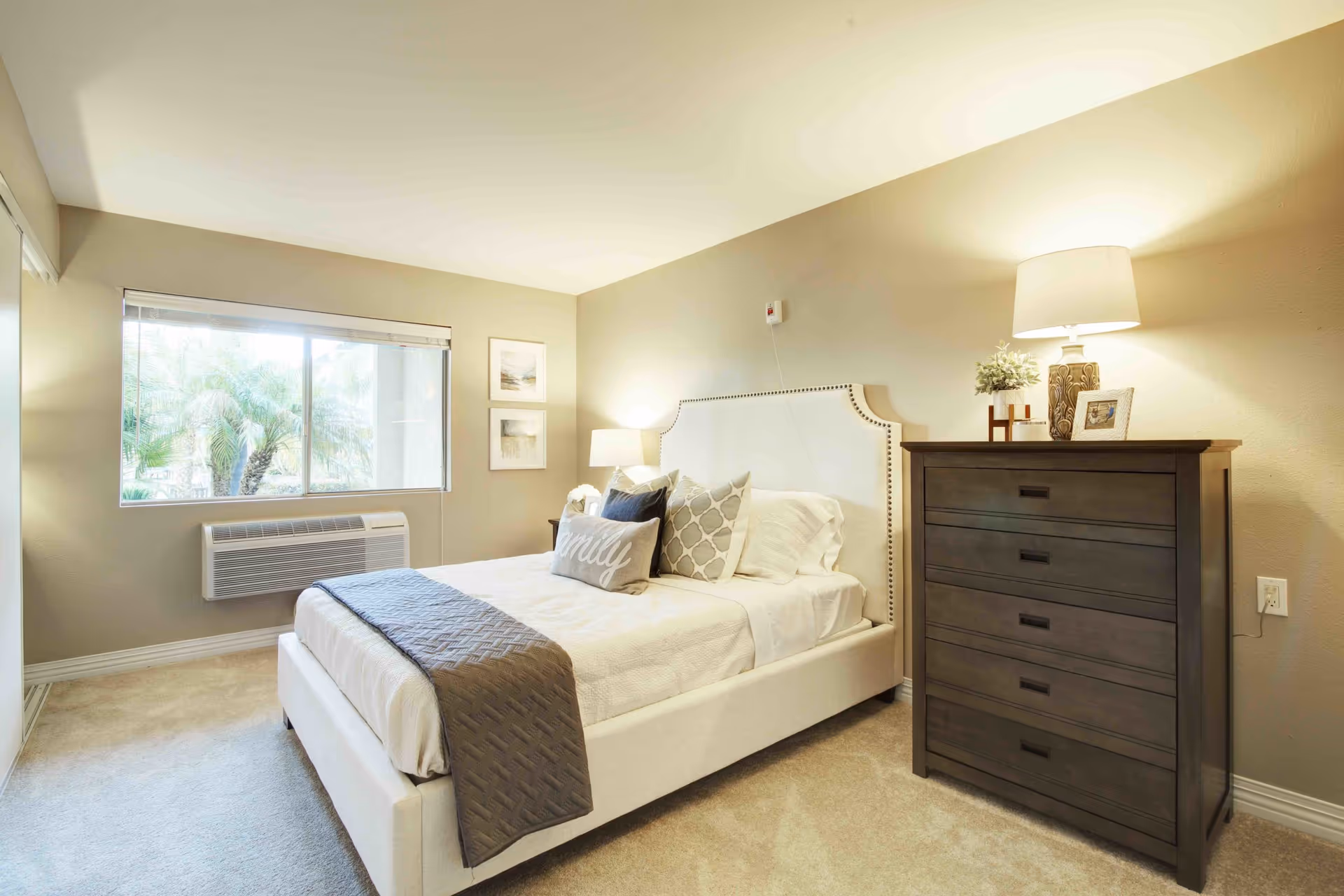 A bright and cozy bedroom with a large window showing palm trees outside. The room features a white upholstered bed with decorative pillows and a gray throw blanket. There is a dark wooden dresser with a lamp, a small plant, and a framed photo on top. The walls are painted beige, and the carpet is light-colored.