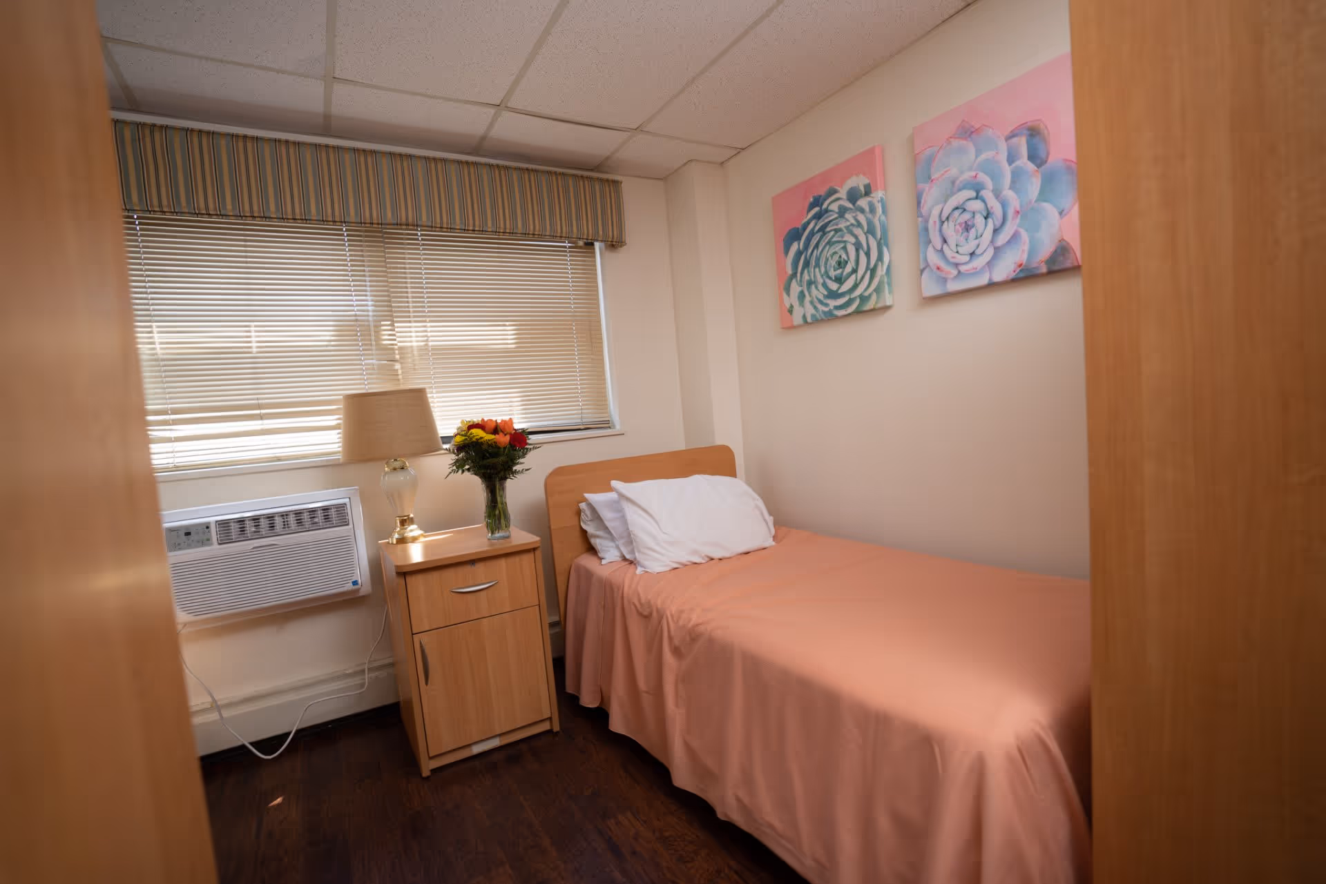 A small bedroom with a single bed covered in a peach-colored bedspread and white pillows. Next to the bed is a wooden nightstand with a lamp and a vase of colorful flowers. The room has a window with closed blinds and a striped valance. Two floral paintings with pink backgrounds hang on the wall above the bed.