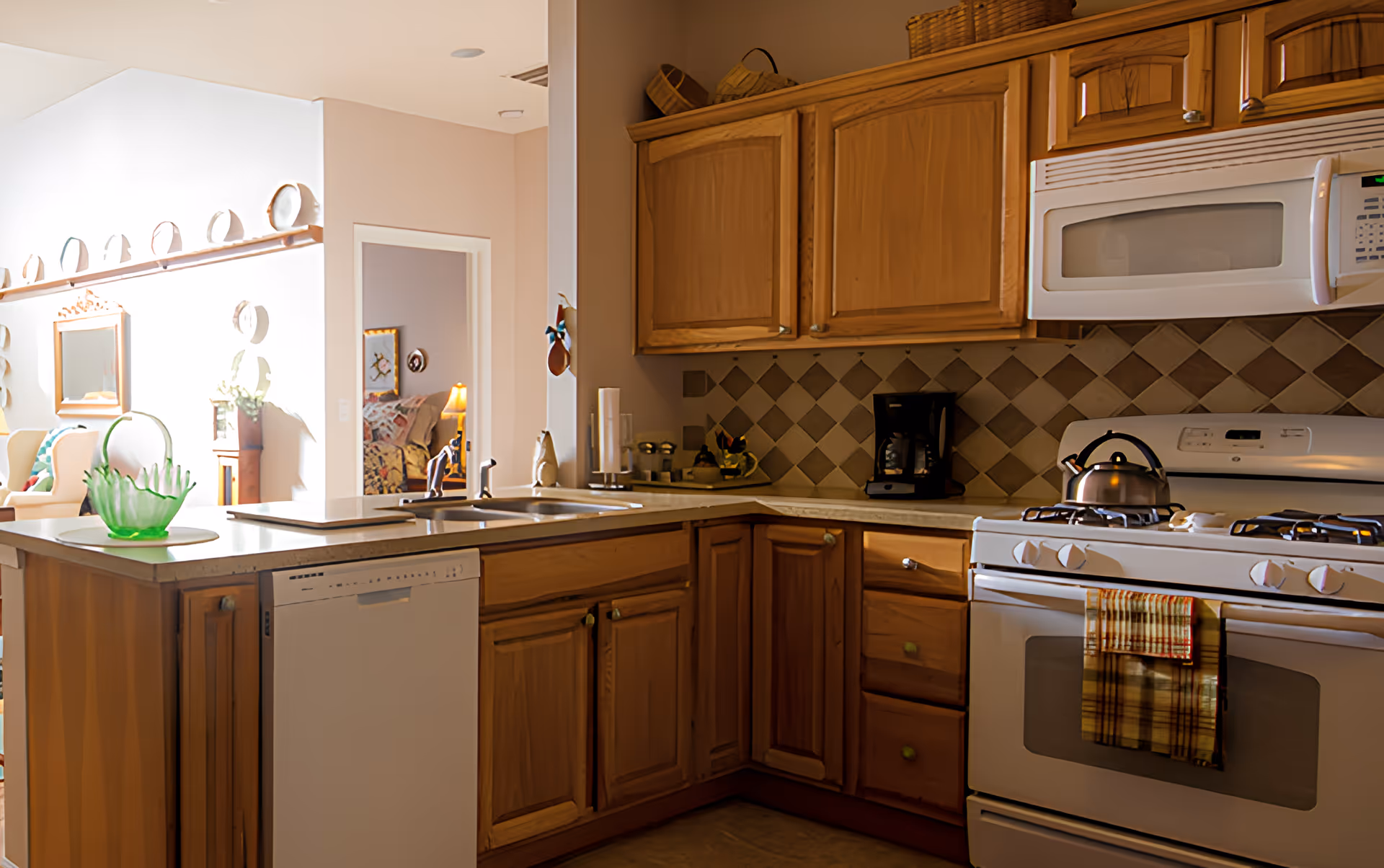 A kitchen area with wooden cabinets, a white dishwasher, a white stove with a kettle on it, and a white microwave above the stove. The countertop has a green decorative glass bowl and a double sink. In the background, there is a glimpse of a living area with chairs, a mirror, and a lamp.