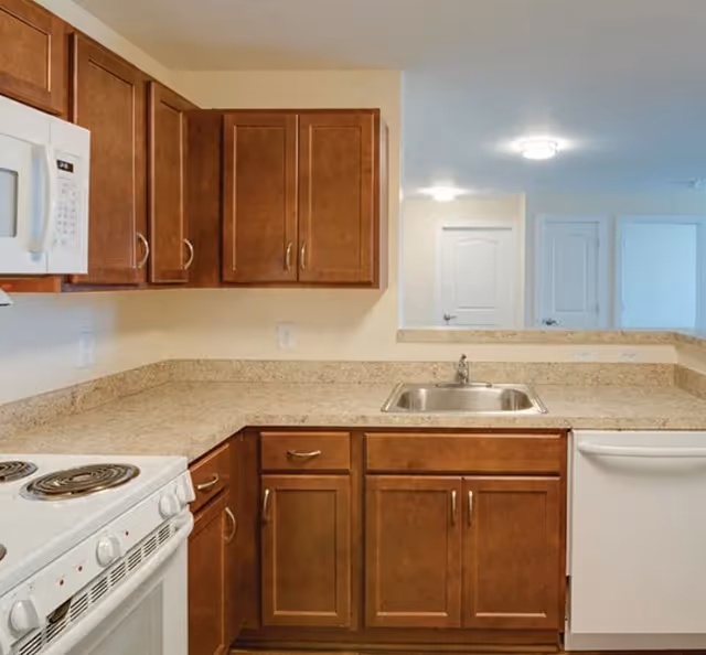 Kitchen with brown wooden cabinets, beige laminate countertops, a stainless sink, white stove and dishwasher, and a pass-through to a hallway.