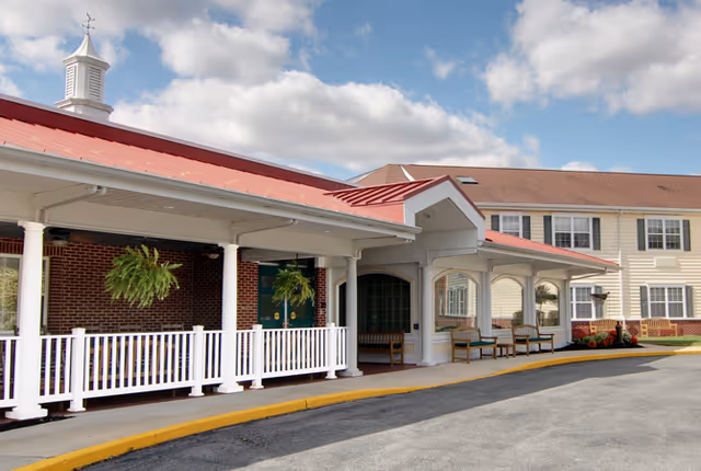 Covered entrance with white columns, hanging ferns and benches at the front of a multi-story senior living building under a partly cloudy sky.
