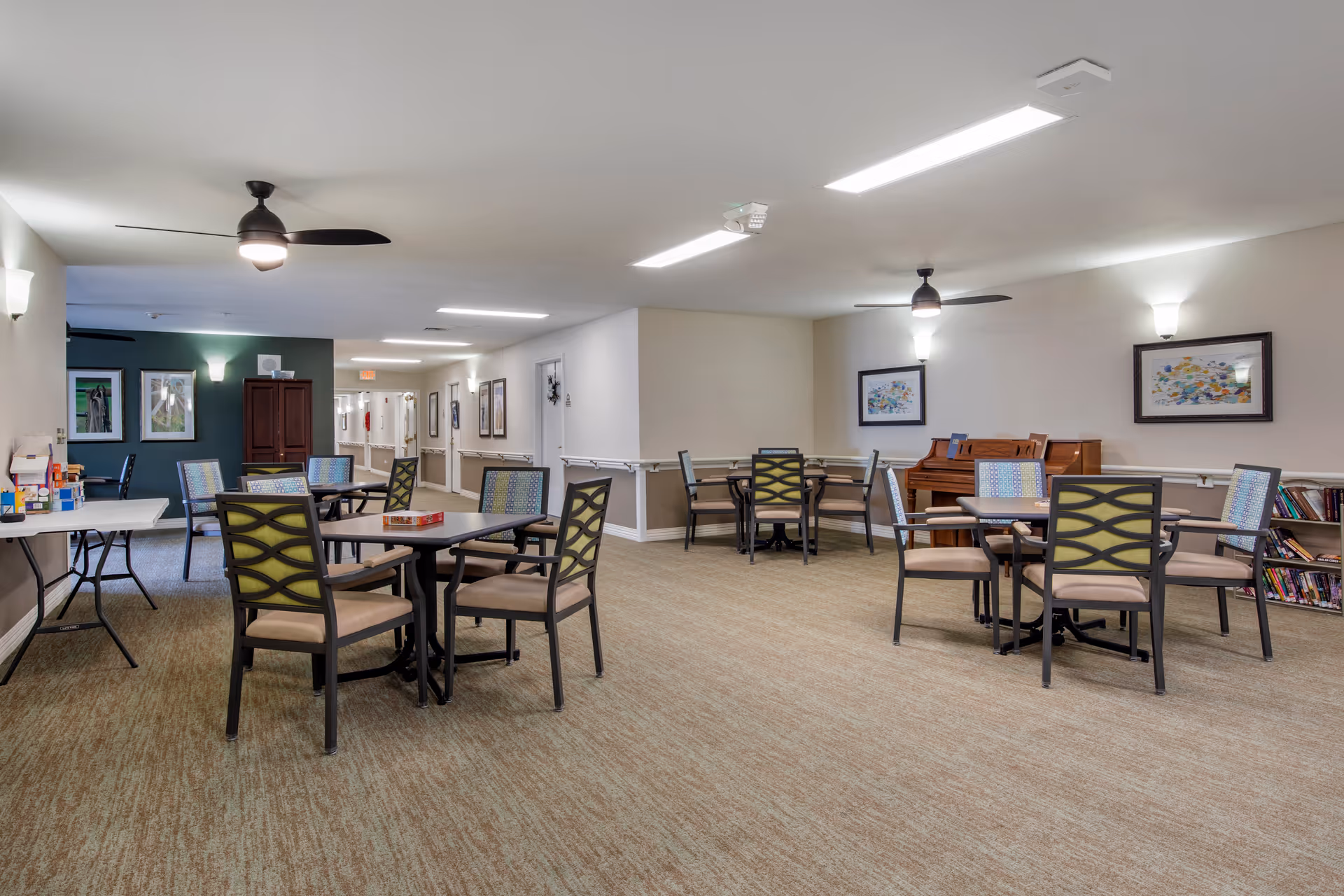 A spacious common area in a senior living facility with several tables and chairs arranged for socializing or activities. The room has beige carpet, white walls with framed artwork, ceiling fans with lights, and a piano against one wall. There is a hallway visible in the background with additional framed pictures and wall-mounted lights.