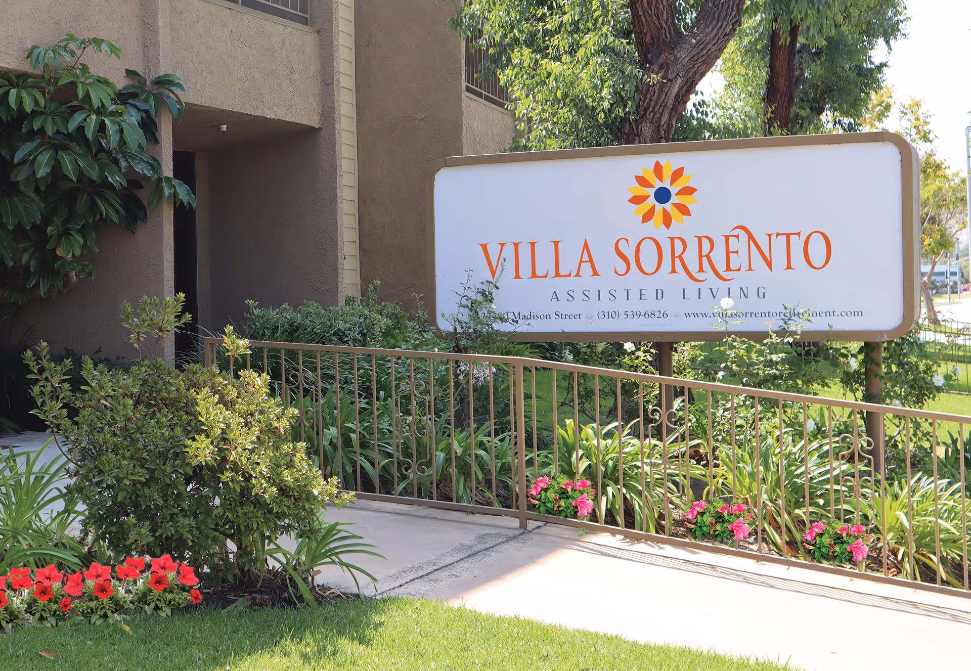 Outdoor view of the entrance area to Villa Sorrento Assisted Living facility, featuring a large sign with the facility's name and logo, surrounded by green plants, flowers, and a metal railing.